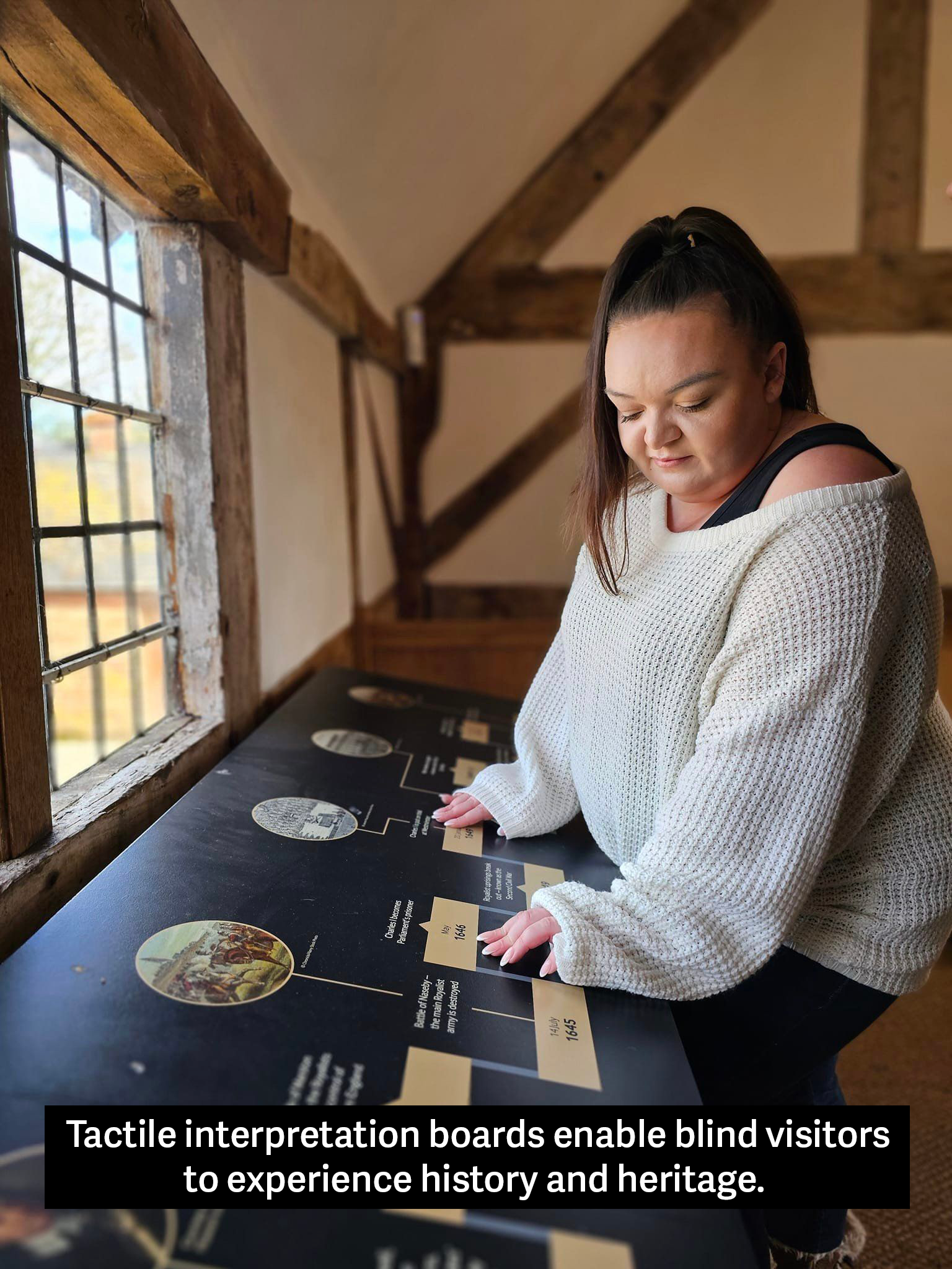 Image depicts a person using their hands to feel along an interpretation board which is situated in front of an old-looking window with light coming into the historic space.