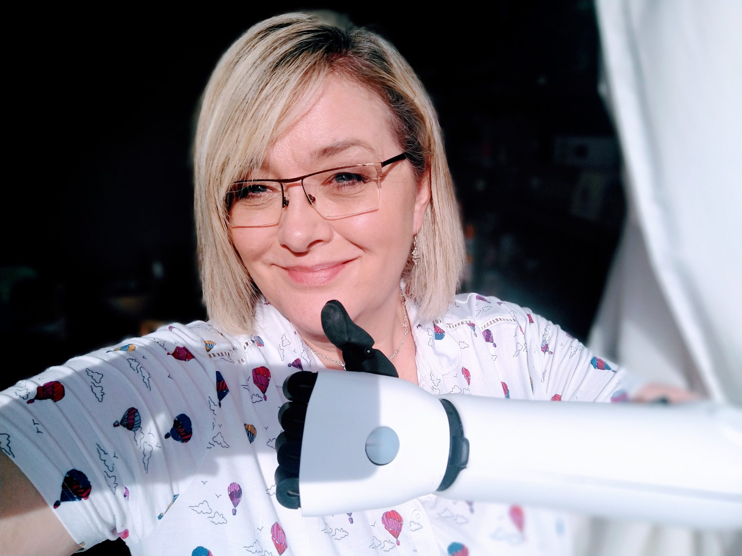 A blonde woman wearing glasses and a white polo shirt with colourful hot balloon print gives a thumbs up sign with her white bionic arm