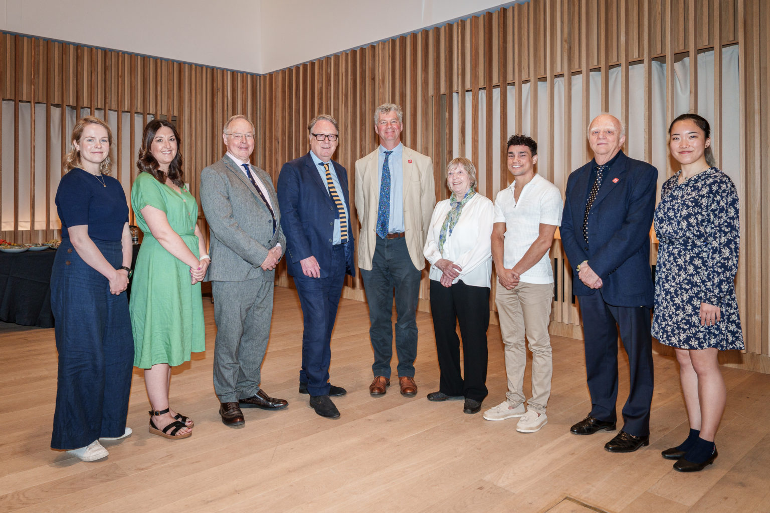 Image shows nine people standing in the line looking towards the camera as a formal pose.  They are indoors standing against a modern interior with light wooden floors and wall panels.  The people are a diverse group and all look happy to be there.