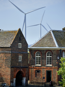 Image shows historic buildings in the foreground and due to perspective large wind turbines immediately behind, against the backdrop of a blue sky